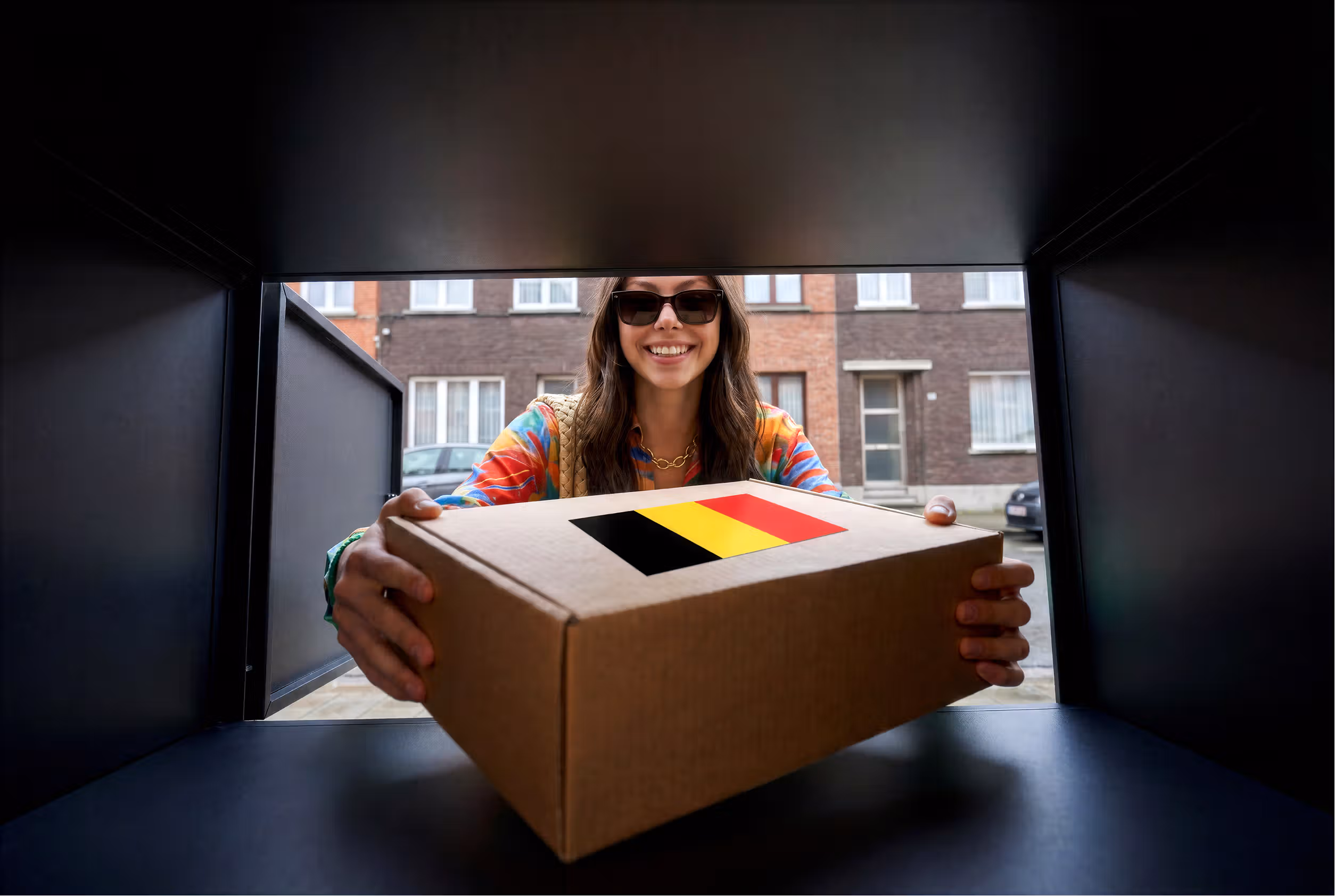Mulher sorridente com óculos de sol entrega uma caixa de cartão através de um Locker ou compartimento de correio. A caixa tem um autocolante com a bandeira da Bélgica.
